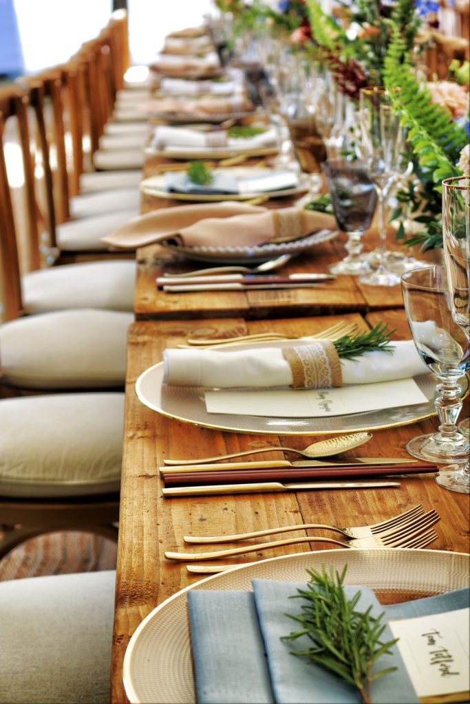 Long wooden dinner table set for a party with gold silverware, cloth napkins, and rosemary sprigs